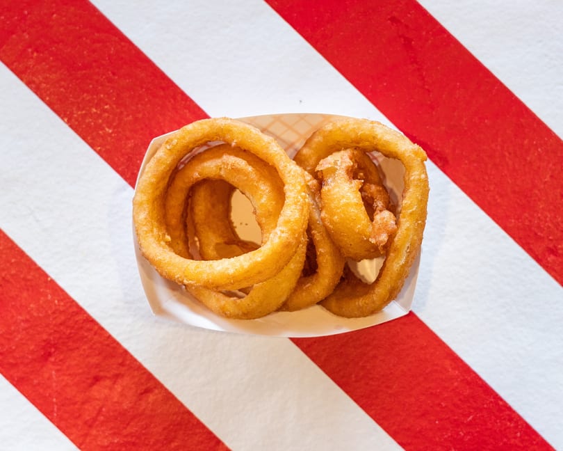 Golden-brown onion rings in a white paper tray, centered on a red and white striped background.
