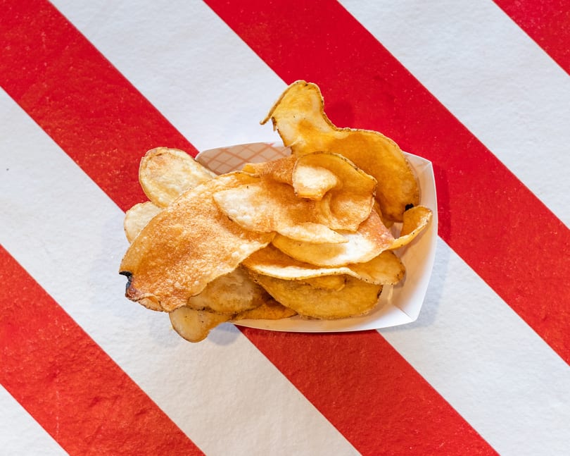 Crispy golden potato chips in a white paper tray, centered on a red and white striped surface.