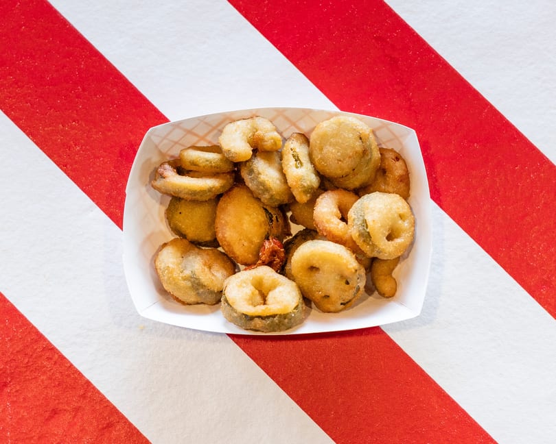 A white paper tray filled with golden-brown fried pickles, some round, some sliced, on a red and white striped surface.