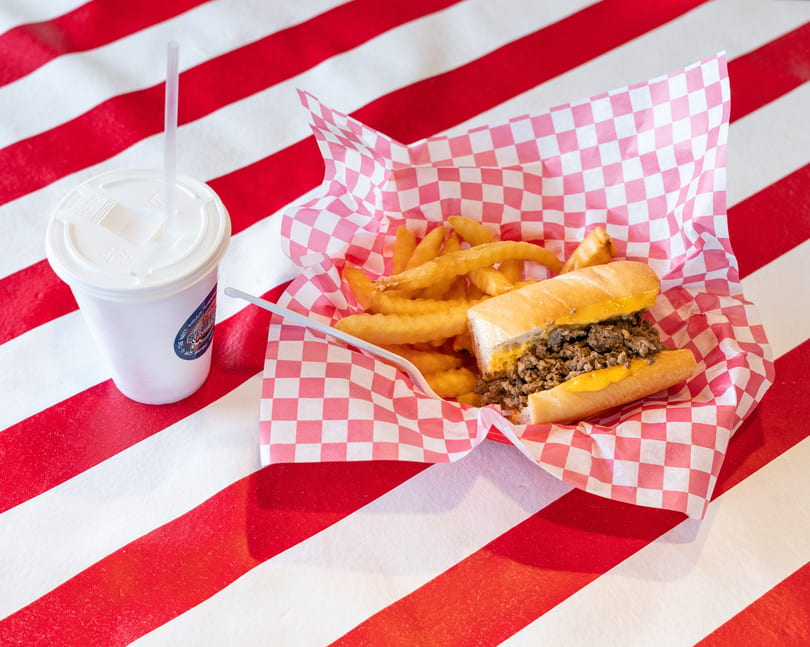 Cheesesteak sandwich and fries in a red checkered basket with a drink on a striped table.