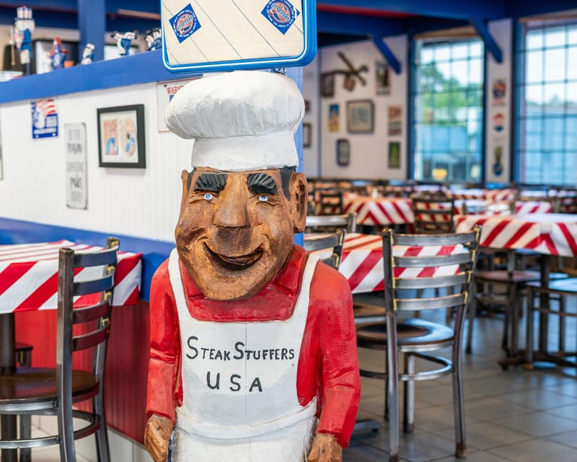 A wooden chef statue with "Steak Stuffers USA" on its apron stands in a restaurant dining room.