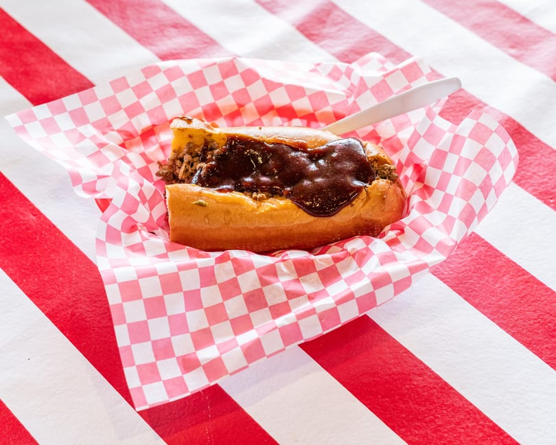 A chili dog with dark sauce and ground meat in a red and white checkered paper boat.