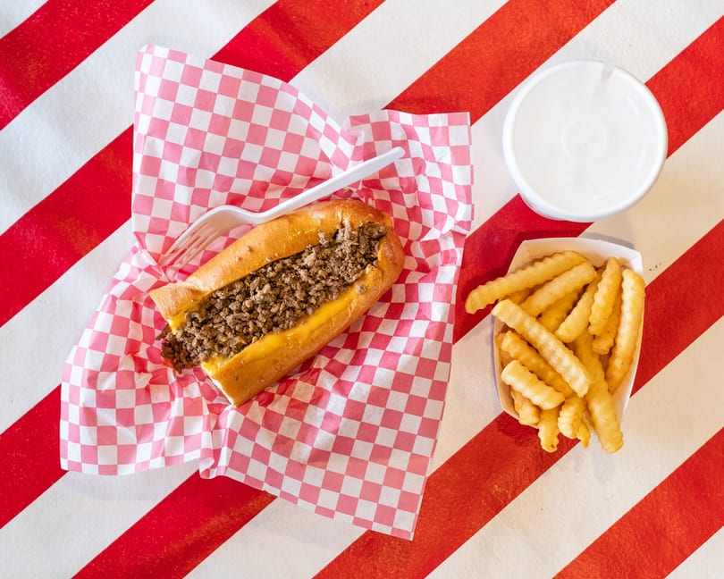 Cheesesteak sandwich with fries and a drink on a red and white striped background.