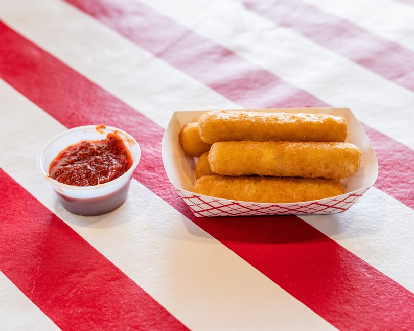 Golden-brown mozzarella sticks in a paper tray with a side of marinara sauce on a red and white striped table.
