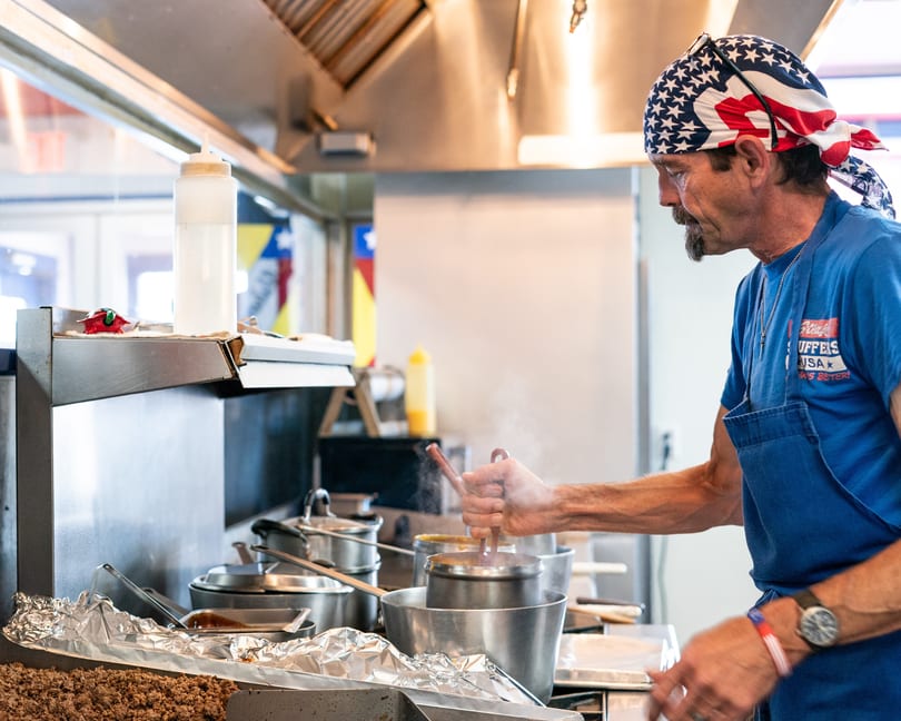 A chef in an American flag bandana stirs a steaming pot in a busy commercial kitchen.