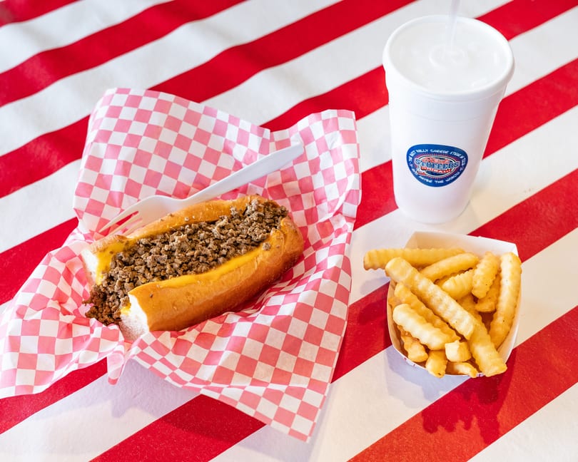 Cheesesteak in checkered paper, crinkle-cut fries, and a drink on a red and white striped table.
