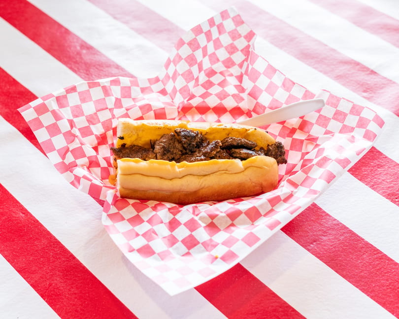 Cheesesteak sandwich with melted cheese and steak in a red and white checkered wrapper on a striped table.