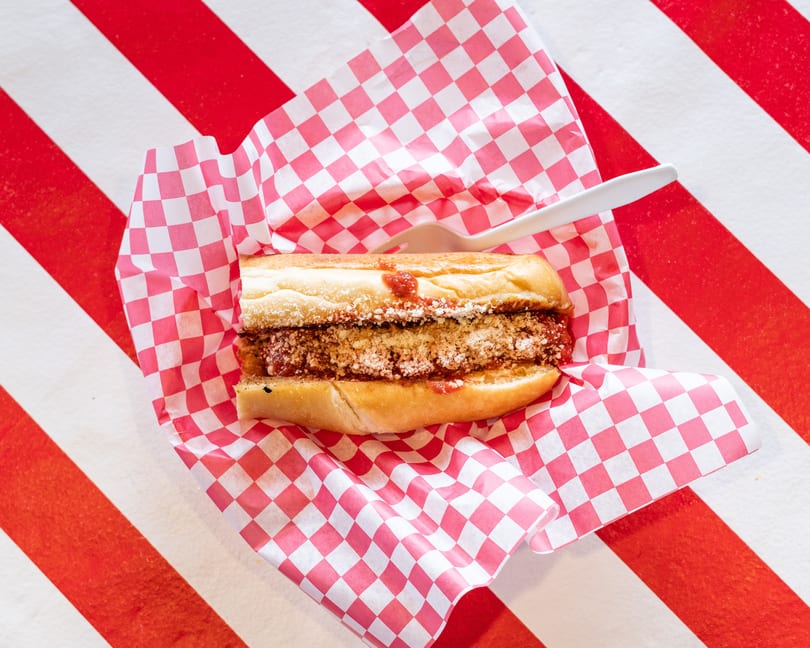 Meatball sub sandwich with sauce and cheese in red and white checkered paper on a striped table.