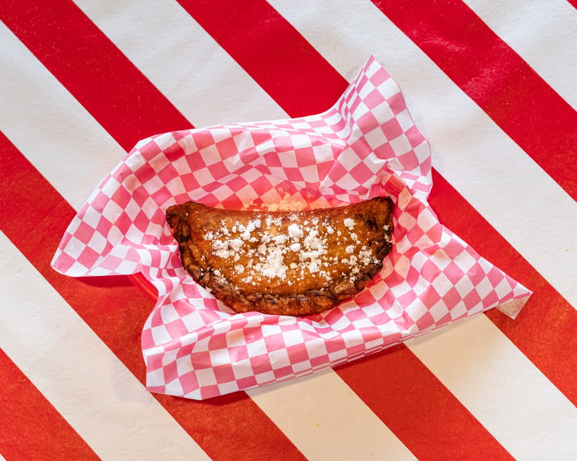 A golden-brown fried dessert empanada, dusted with powdered sugar, rests in a pink checkered paper.